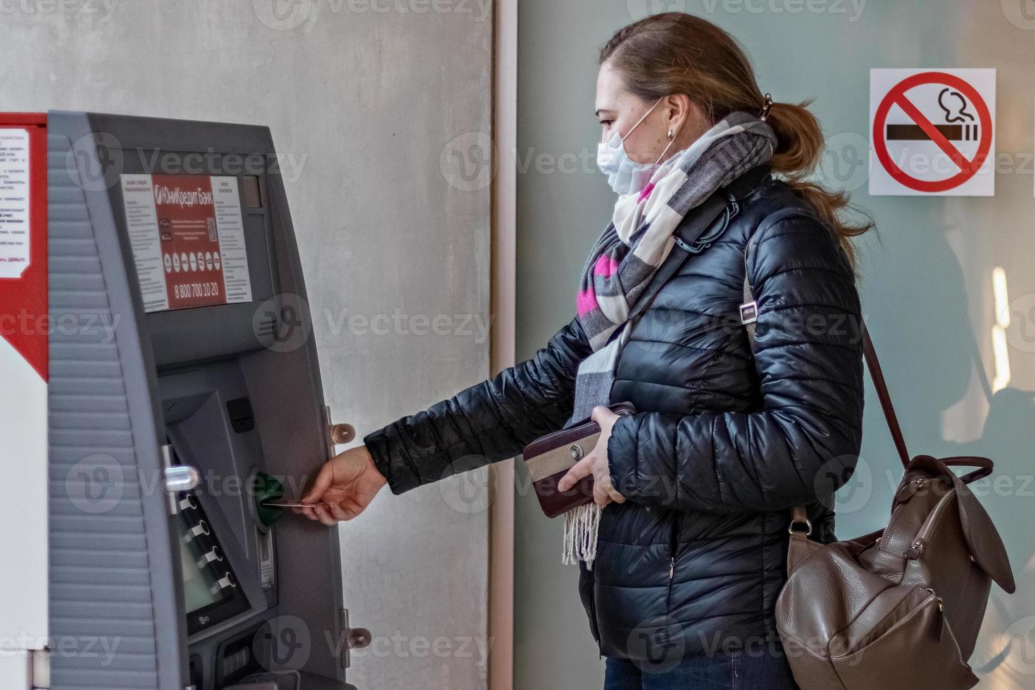 een vrouw die een beschermend medisch masker op haar gezicht draagt, een creditcard invoert en geld opneemt bij een geldautomaat. foto