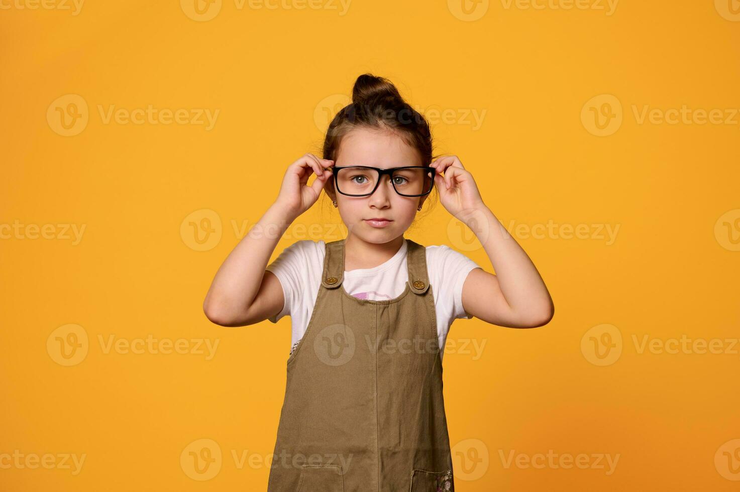 zelfverzekerd 6 jaren oud weinig kind meisje, primair school- leerling, vervelend bril, op zoek Bij camera, oranje achtergrond foto