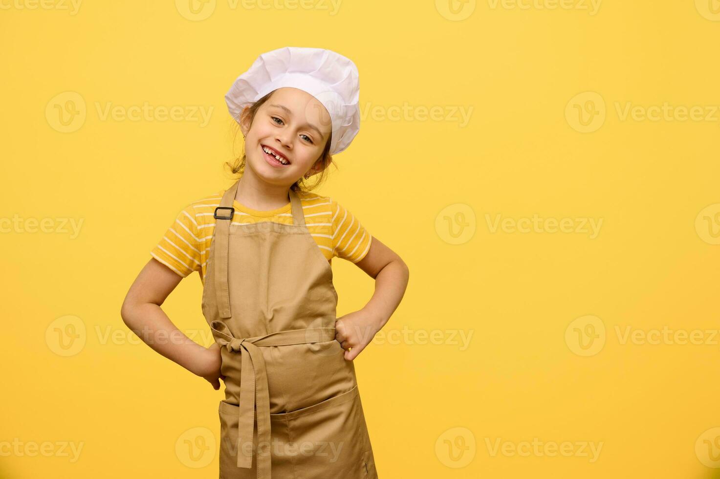 schattig kind meisje, weinig chef banketbakker in schort en wit chef pet, zetten handen Aan taille, glimlachen op zoek Bij camera foto