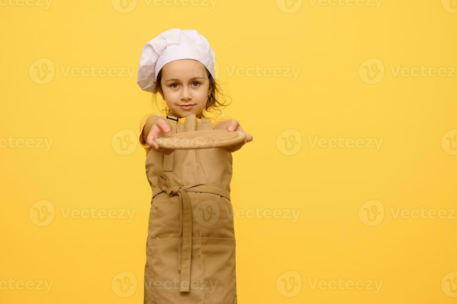 weinig meisje in chef hoed en schort, glimlacht en houdt uit Bij camera een houten bord, geïsoleerd over- geel studio achtergrond foto