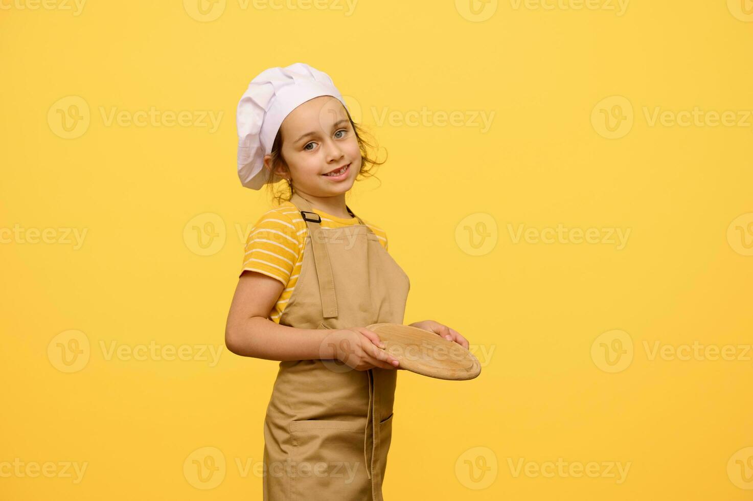 schattig weinig chef banketbakker, koken, bakker, pizzaiolo, aanbiddelijk kind meisje houdt een houten bord, glimlacht Bij camera foto