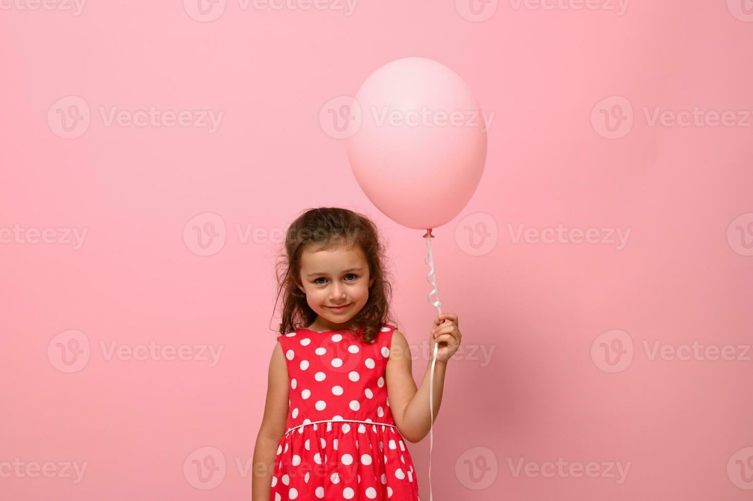 portret van mooi mooi prachtig aanbiddelijk 4 jaren verjaardag meisje, kind in jurk met polka stippen patroon, Holding een roze ballon, geïsoleerd over- roze achtergrond met kopiëren ruimte voor reclame. foto