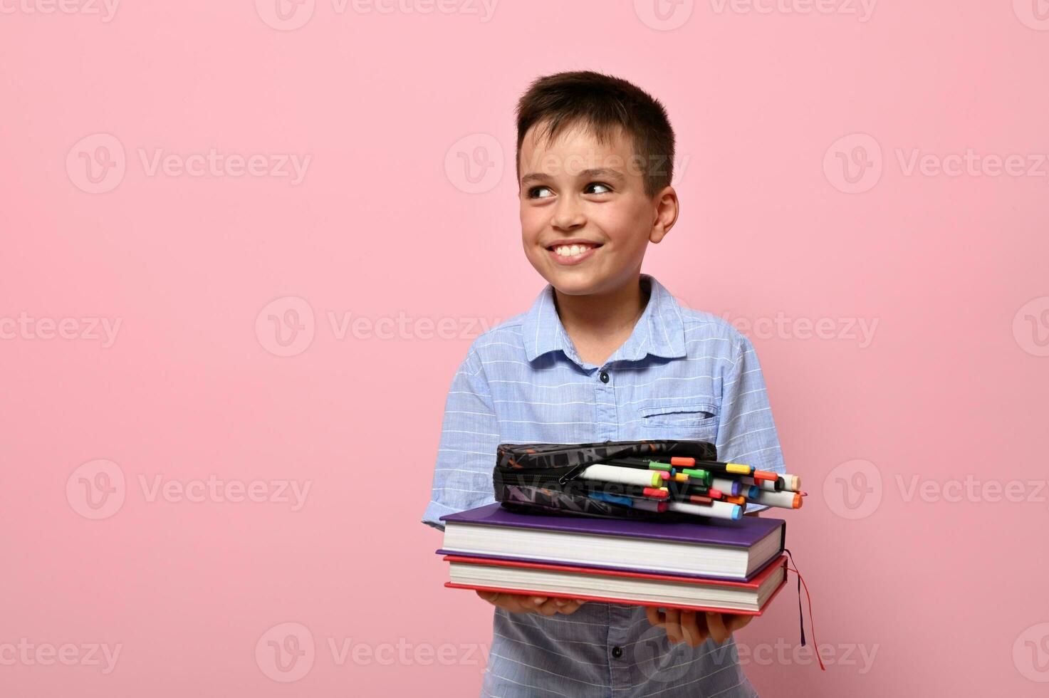 een charmant jongen, een leerling Bij school, houdt boeken in voorkant van hem en potlood geval met pennen, viltstift pennen en markeringen vallend uit van hem. school- jongen schattig glimlacht op zoek naar de kant Aan roze achtergrond foto