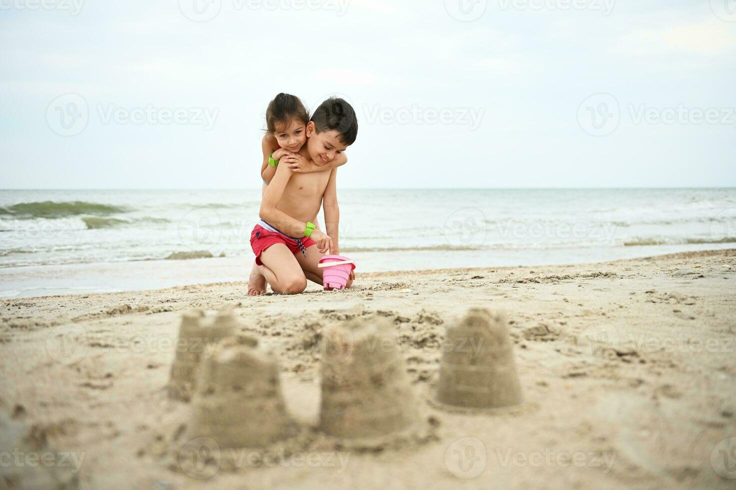 knap jongen vult emmer met zee zand terwijl zijn jonger zus voorzichtig knuffels hem. mooi meisje knuffels haar broer schattig terwijl spelen strand spellen. wazig gebouwd zandkastelen in de voorgrond foto