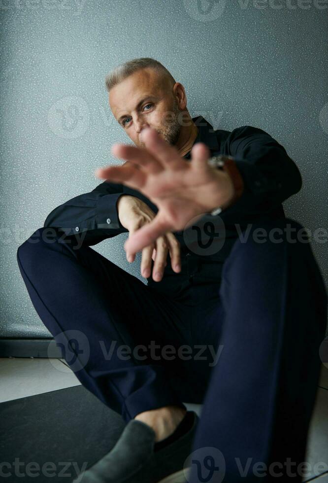 knap aantrekkelijk Kaukasisch geslaagd midden- oud Mens in zwart gewoontjes pak op zoek vol vertrouwen Bij camera Holding palm uit naar camera poseren tegen grijs muur foto