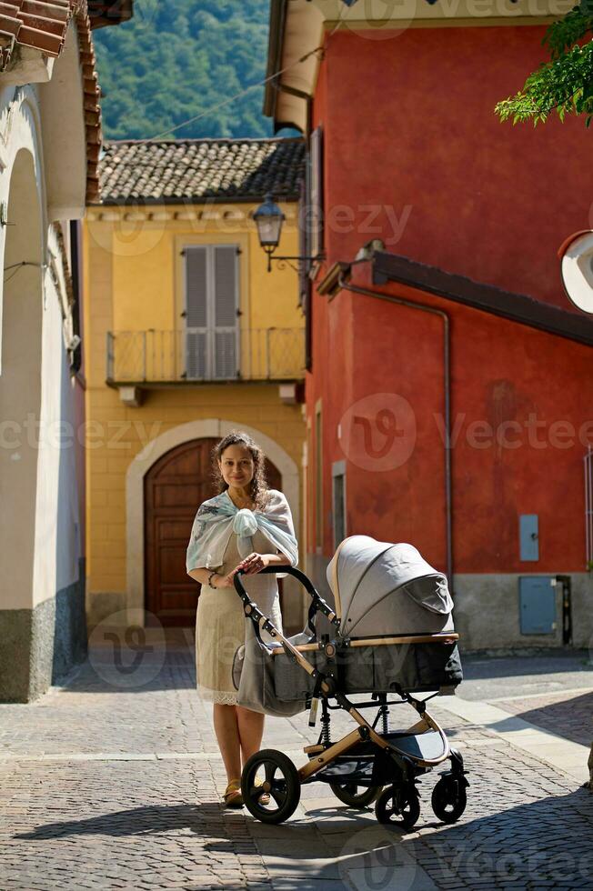 jong moeder op zoek Bij camera, wandelen langs geplaveide steeg in Italiaans middeleeuws dorp, met haar baby slapen in kinderwagen foto