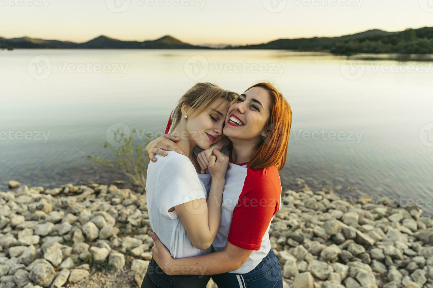 gelukkig vrouw omarmen vriendin terwijl staand door meer gedurende zonsondergang foto