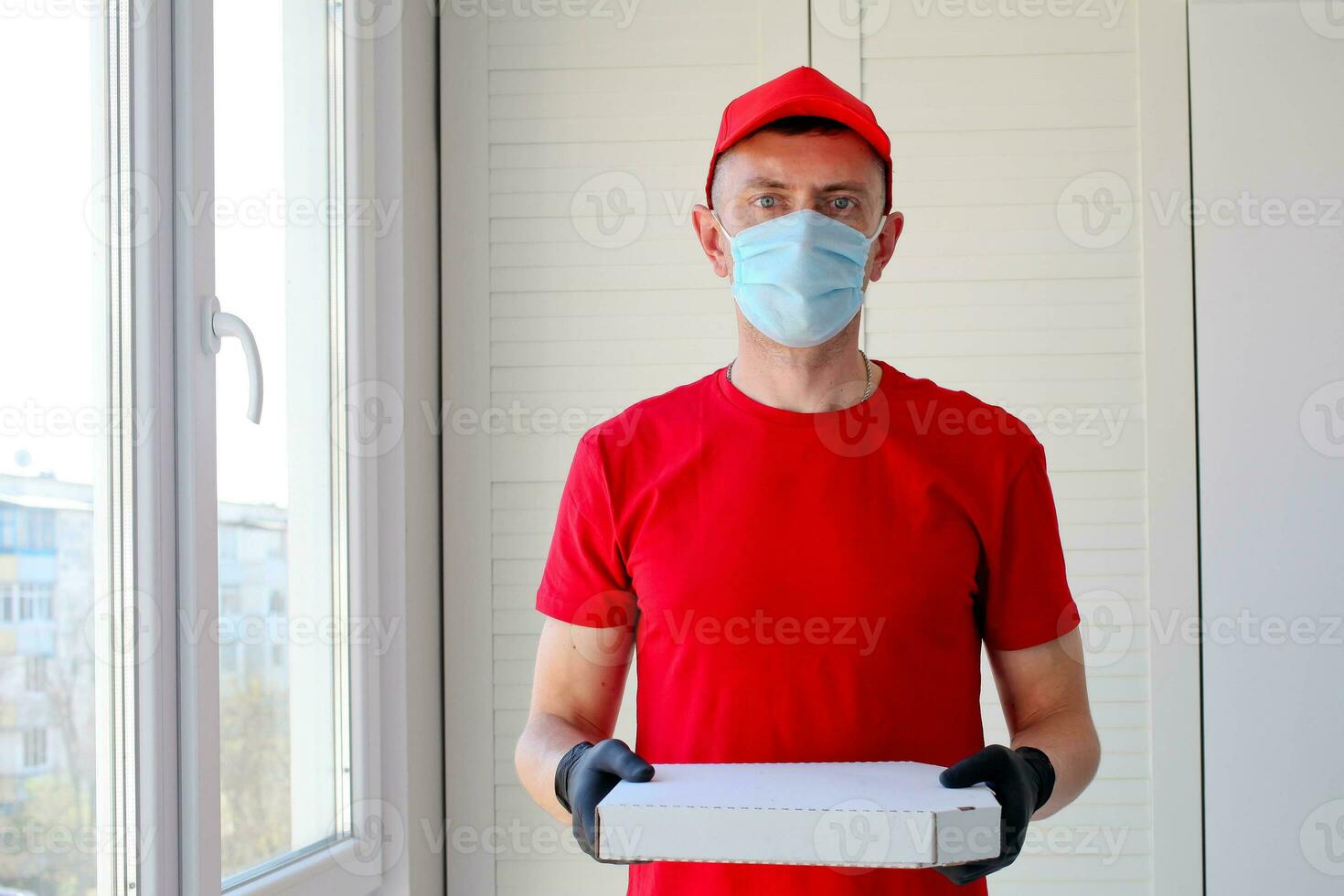 levering Mens werknemer in rood pet blanco t-shirt uniform gezicht masker handschoenen houden karton doos pizza . onderhoud quarantaine pandemisch coronavirus virus 2019-ncov concept. foto