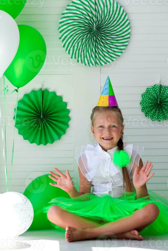 weinig meisje met een pet Aan haar hoofd tegen de achtergrond van groen ballonnen. verjaardag voor kinderen. foto