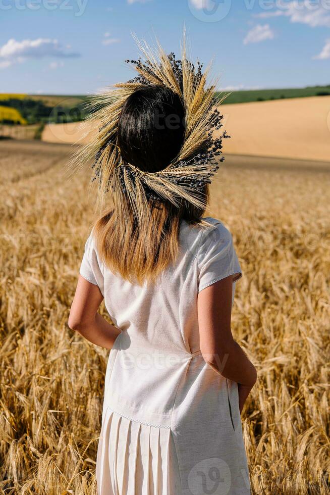 vrouw in krans van bloemen met tarwe en wit jurk wandelen langs ontbijtgranen veld- foto