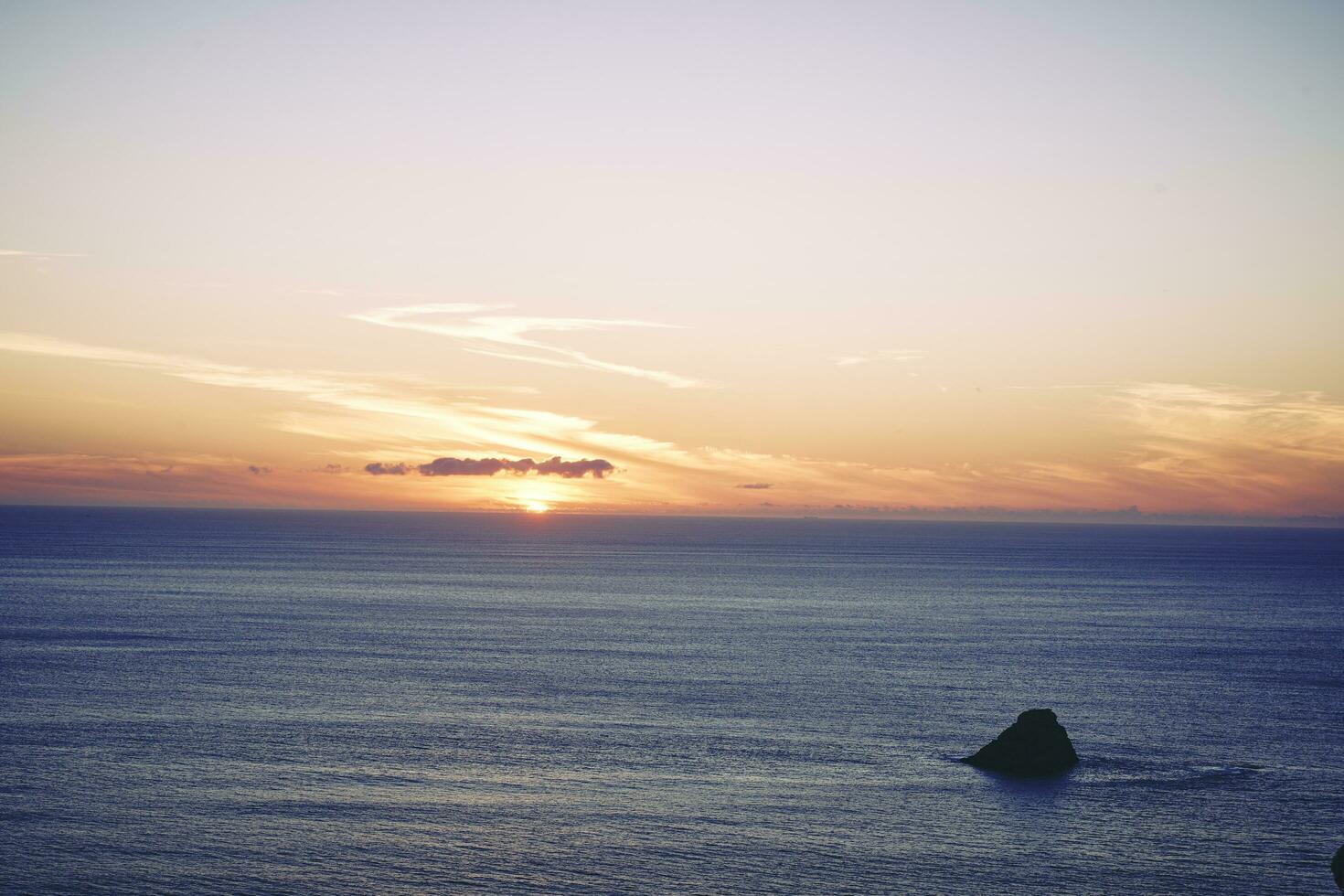 kust- kalmte Bij zonsondergang. prominent rots eiland in bijna kalmte zee onder een gouden bewolkt lucht foto