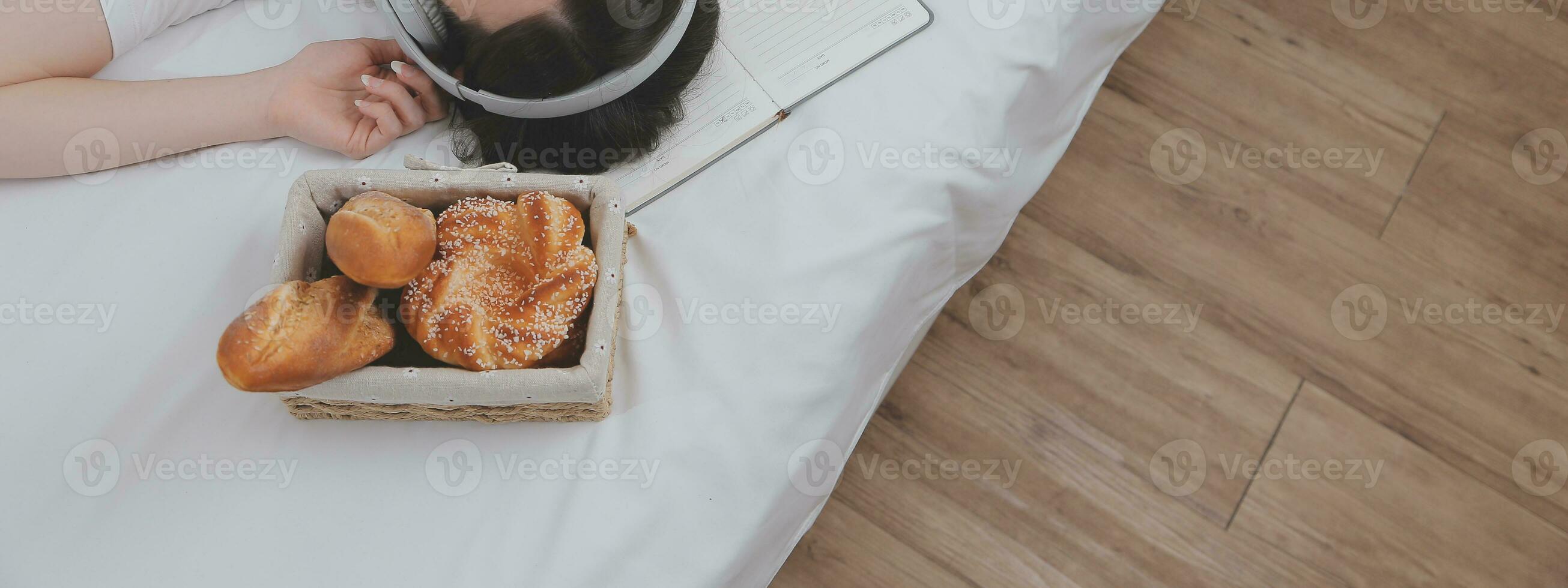 doeltreffend jong millennial meisje zittend Aan een bed in de ochtend, toepassingen laptop computer en eet croissants en drankjes koffie voor ontbijt foto