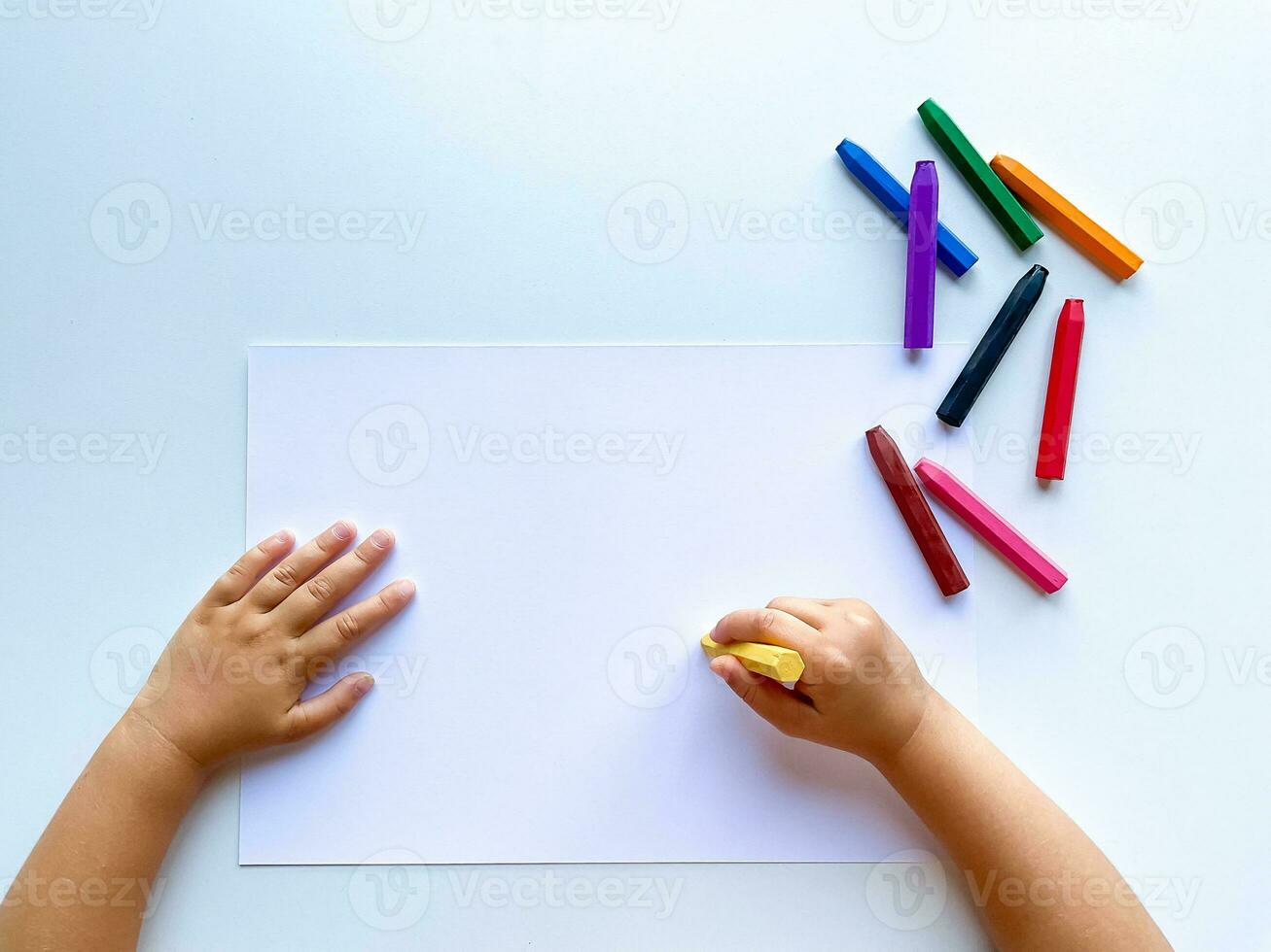 kinderen handen trek met gekleurde was- kleurpotloden Aan een wit vel van papier. top visie van een blanco vel. foto