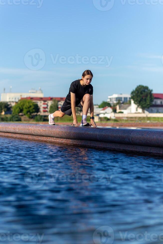 jong, fit en sportief meisje in zwart kleren uitrekken na de training in de buurt de openbaar fontein in de stedelijk stad park. foto