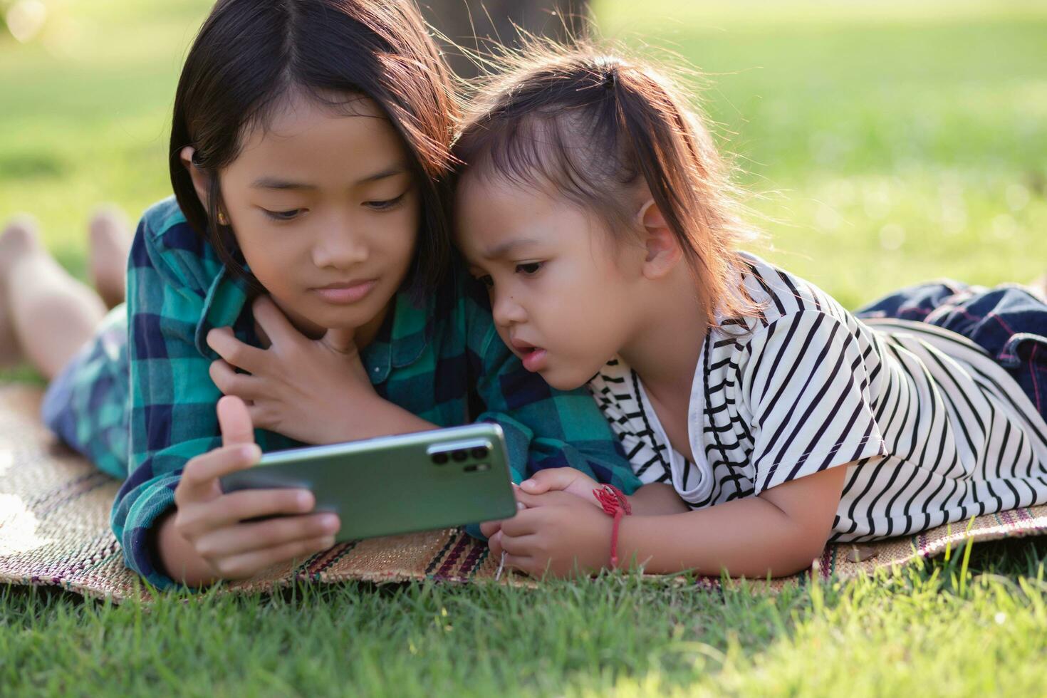 twee jong meisjes aan het liegen Aan de gazon op zoek Bij hun telefoons, zomer, gouden uur, zonsondergang. sstkhuis foto