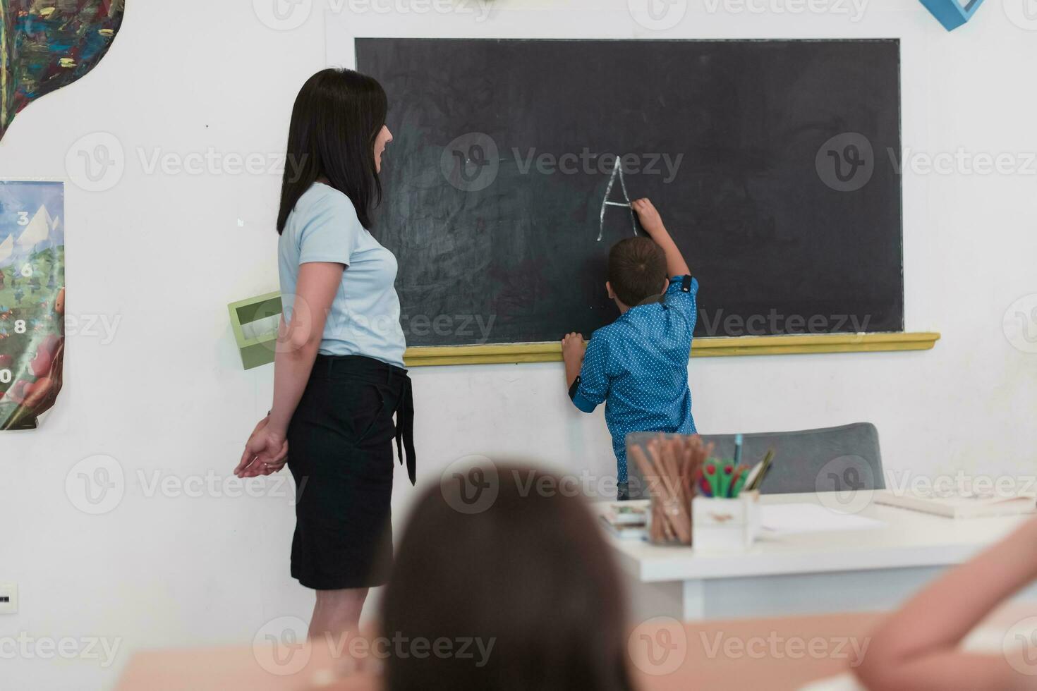 elementair school. de vrouw leraar helpen de kind leerling terwijl schrijven de antwoord Aan de schoolbord. foto