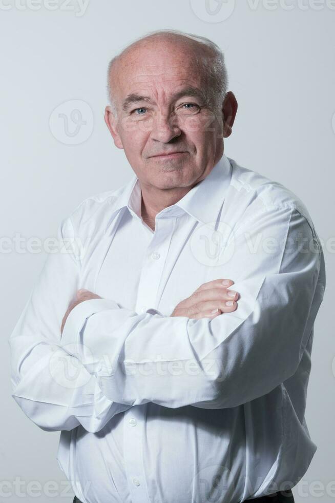 zelfverzekerd senior Mens in wit overhemd kruispunt handen Aan borst en op zoek Bij camera terwijl staand tegen grijs achtergrond. zelf zelfverzekerd senior geïsoleerd wit studio schieten. foto