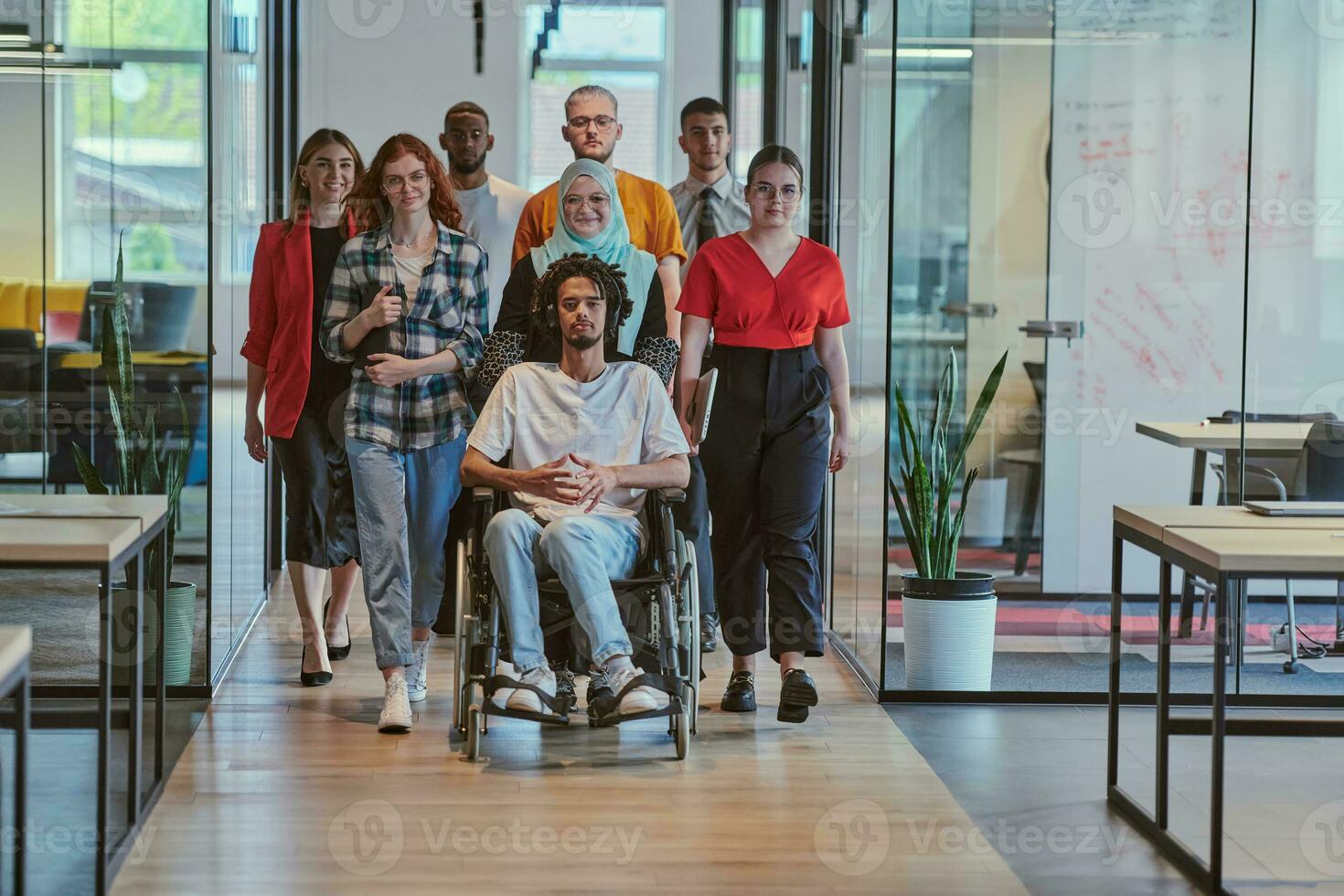 een verschillend groep van jong bedrijf mensen wandelen een gang in de glas-omsloten kantoor van een modern beginnen, inclusief een persoon in een rolstoel en een vrouw vervelend een hijaab, tonen een dynamisch mengen van innovatie en eenheid. foto