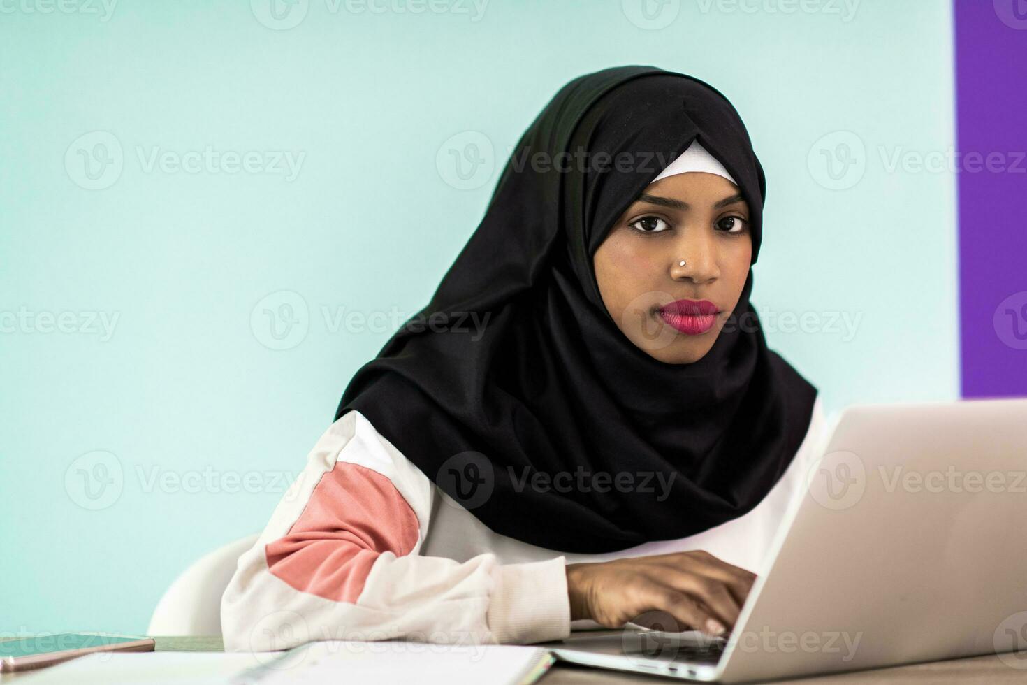 Afrikaanse Amerikaans vrouw vervelend een hijab bedachtzaam zittend in haar huis kantoor en toepassingen een laptop en smartphone foto