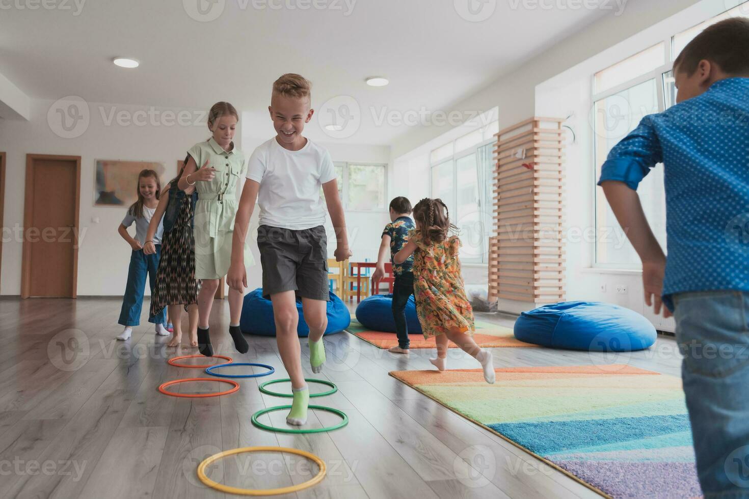 klein kinderkamer school- kinderen met vrouw leraar Aan verdieping binnenshuis in klas, aan het doen oefening. jumping over- hula hoepel cirkels bijhouden Aan de vloer. foto