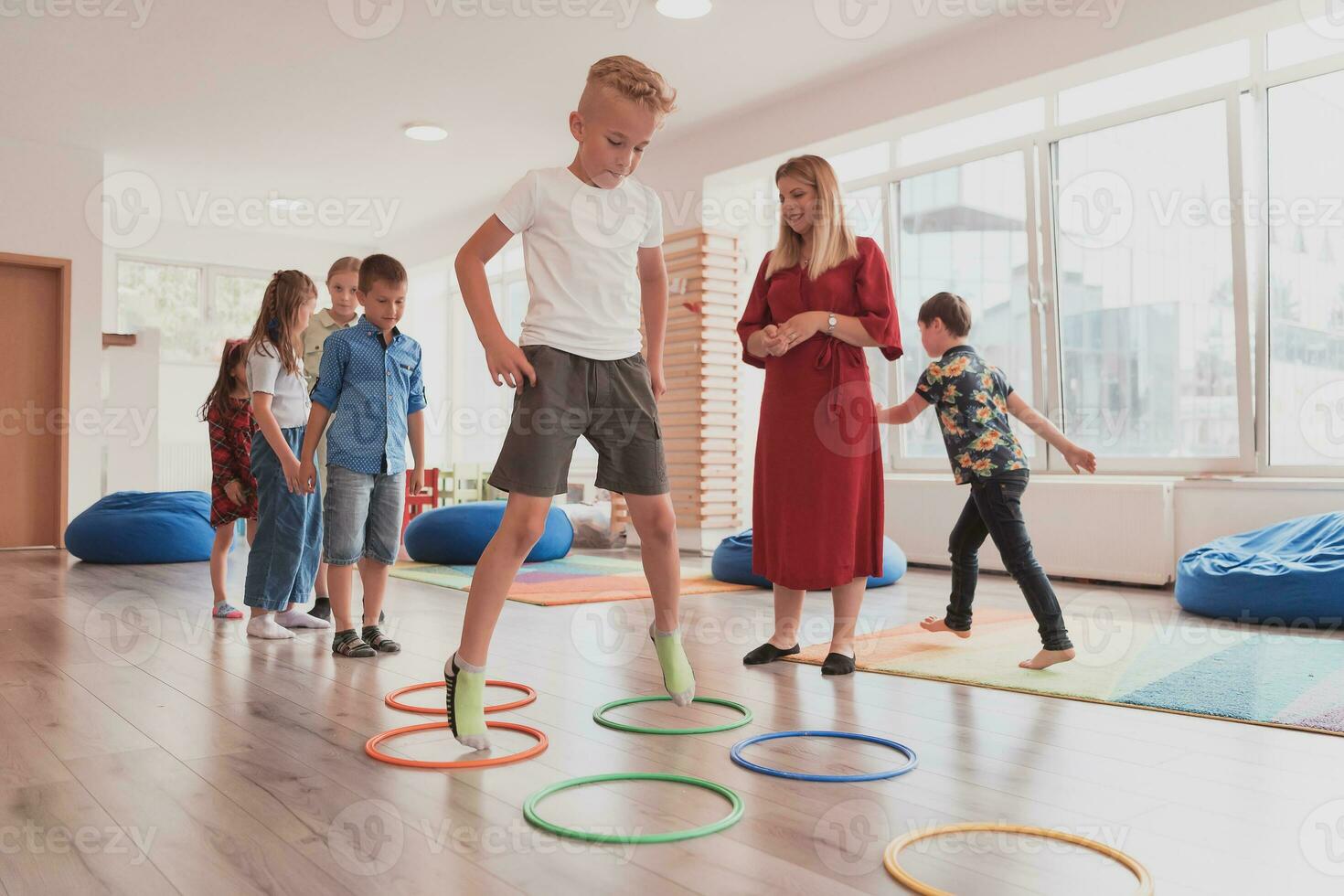 klein kinderkamer school- kinderen met vrouw leraar Aan verdieping binnenshuis in klas, aan het doen oefening. jumping over- hula hoepel cirkels bijhouden Aan de vloer. foto