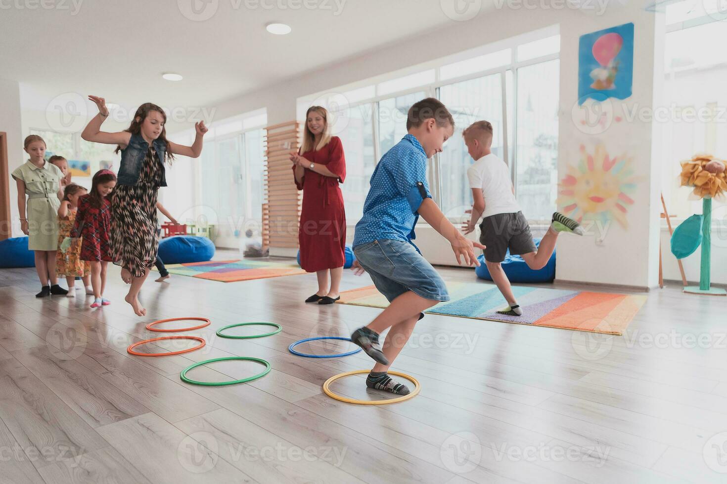 klein kinderkamer school- kinderen met vrouw leraar Aan verdieping binnenshuis in klas, aan het doen oefening. jumping over- hula hoepel cirkels bijhouden Aan de vloer. foto