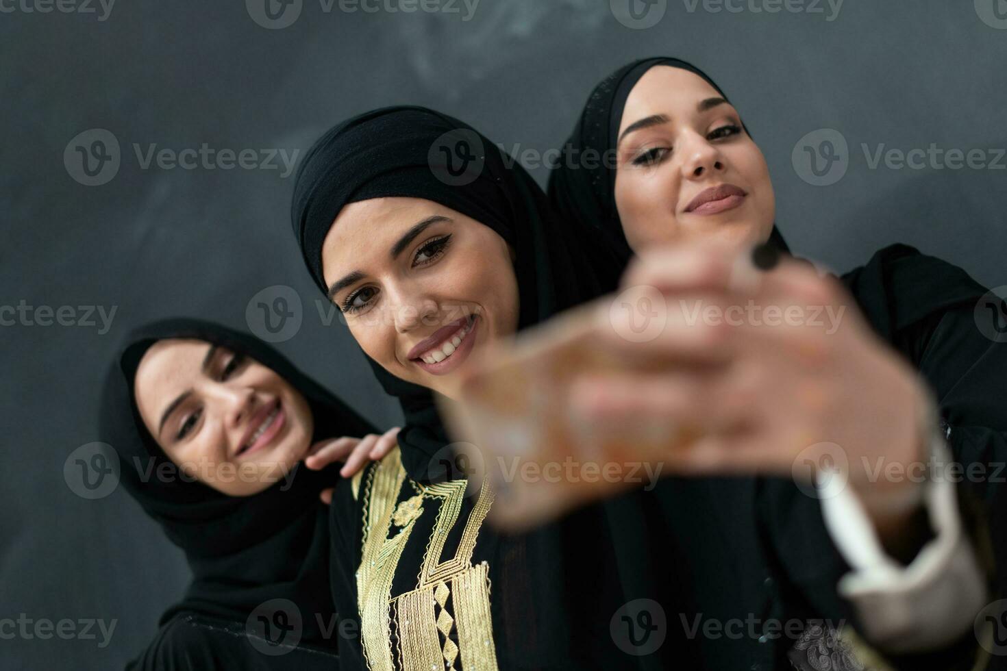 groep van jong mooi moslim Dames in modieus jurk met hijab gebruik makend van smartphone terwijl nemen selfie afbeelding in voorkant van zwart achtergrond foto