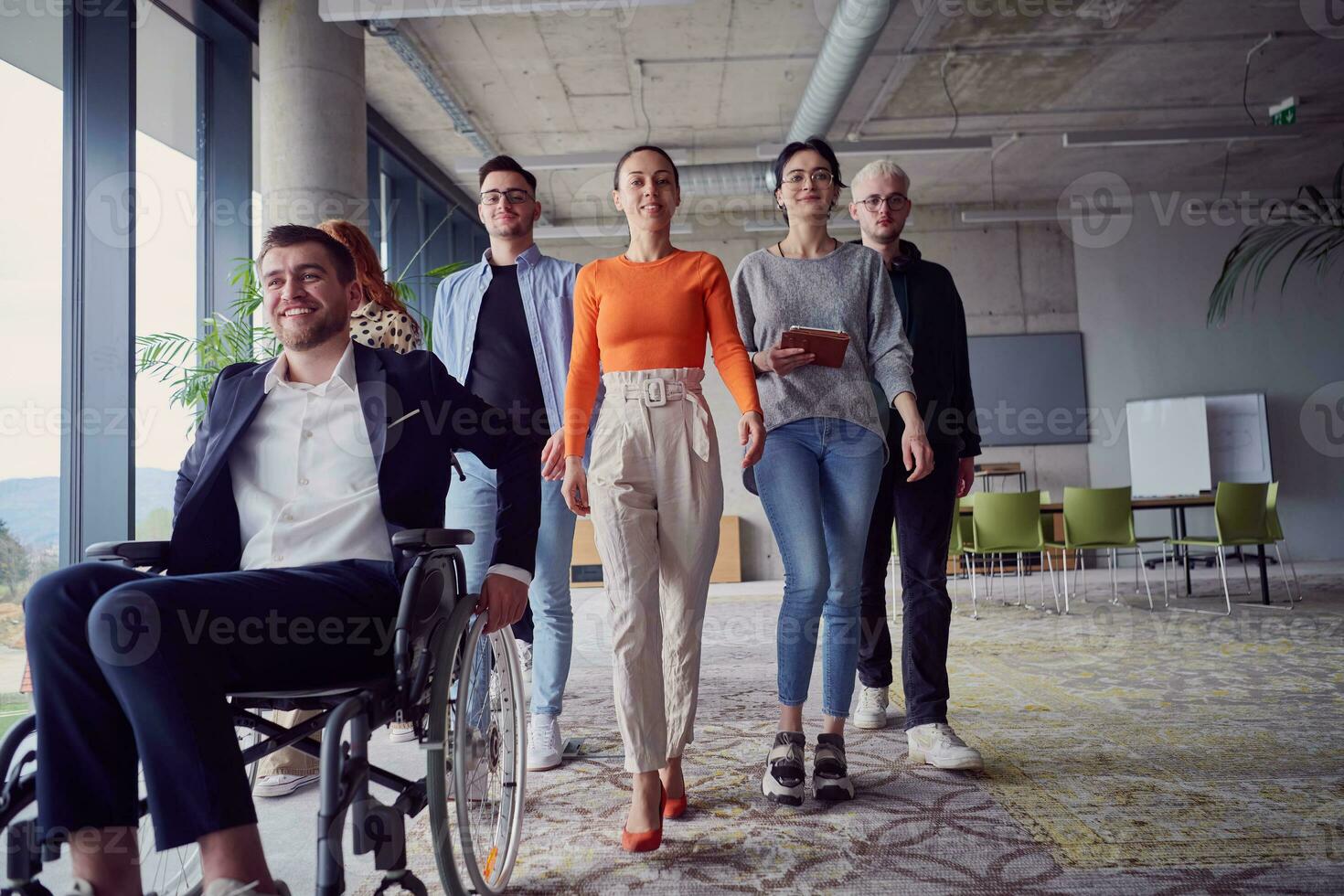een verschillend groep van zakenlieden, inclusief een zakenman in rolstoel, vol vertrouwen pas samen door een modern, ruim kantoor, belichamend samenwerking, inclusiviteit, en sterkte in eenheid foto