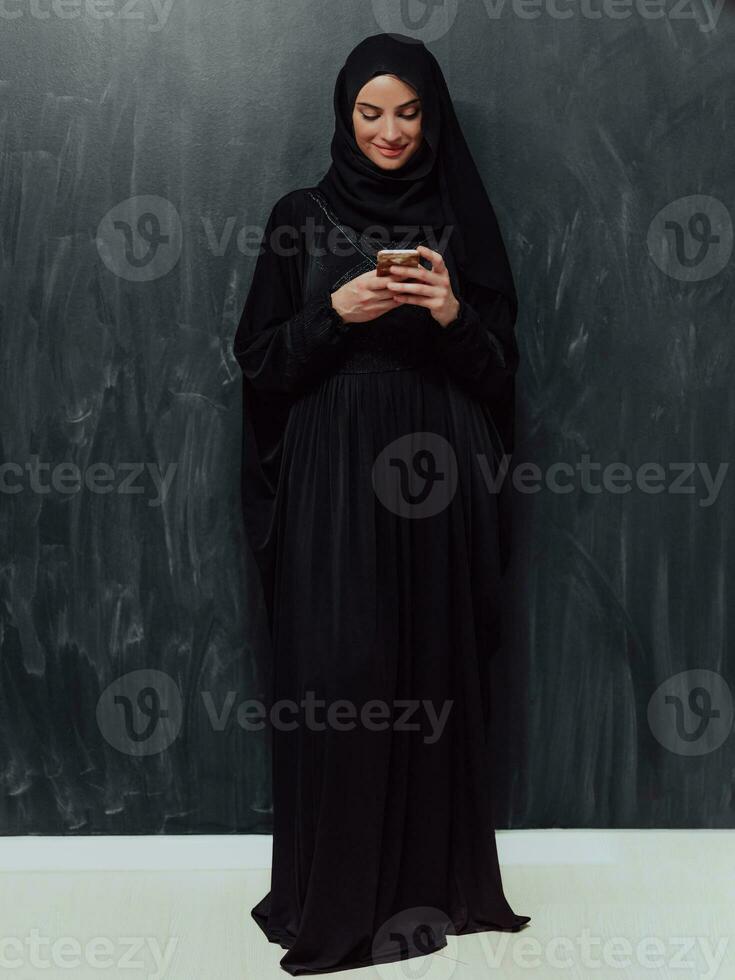 jong modern moslim bedrijf vrouw gebruik makend van smartphone vervelend hijab kleren in voorkant van zwart schoolbord foto