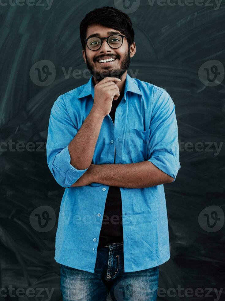 een jong Indisch Mens in een blauw overhemd en bril poses bedachtzaam in voorkant van school- bord foto