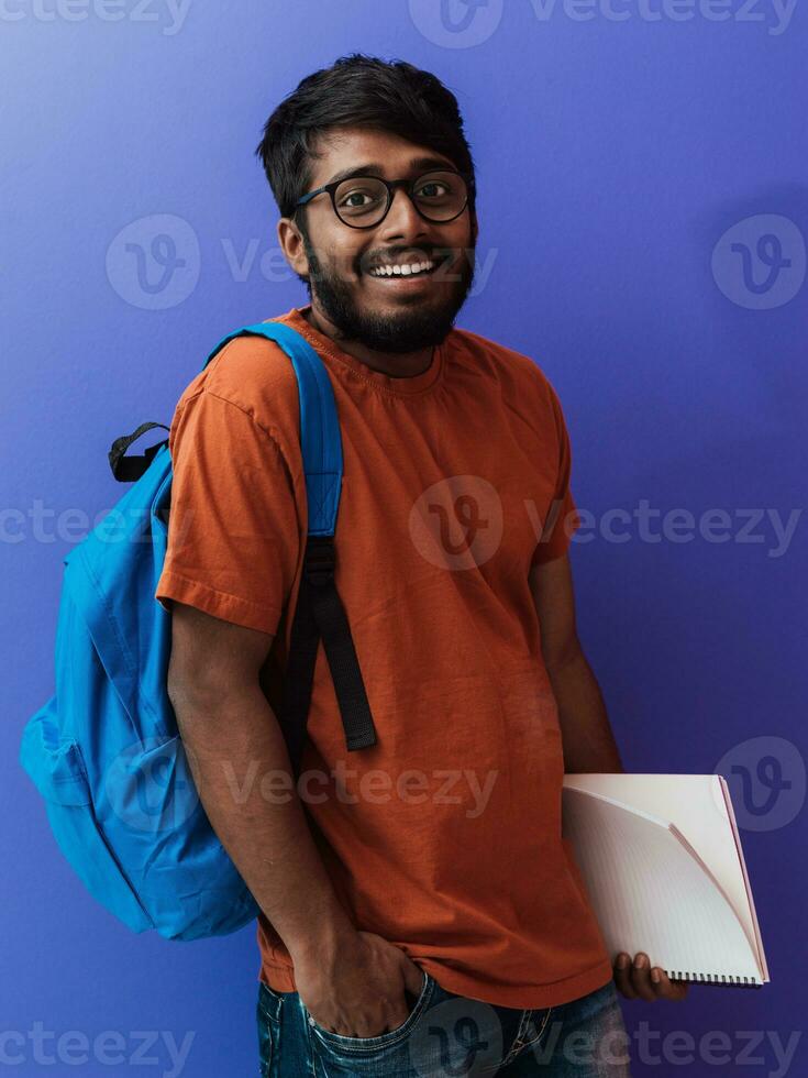 Indisch leerling met blauw rugzak, bril en notitieboekje poseren Aan Purper achtergrond. de concept van onderwijs en scholing. tijd naar Gaan terug naar school- foto