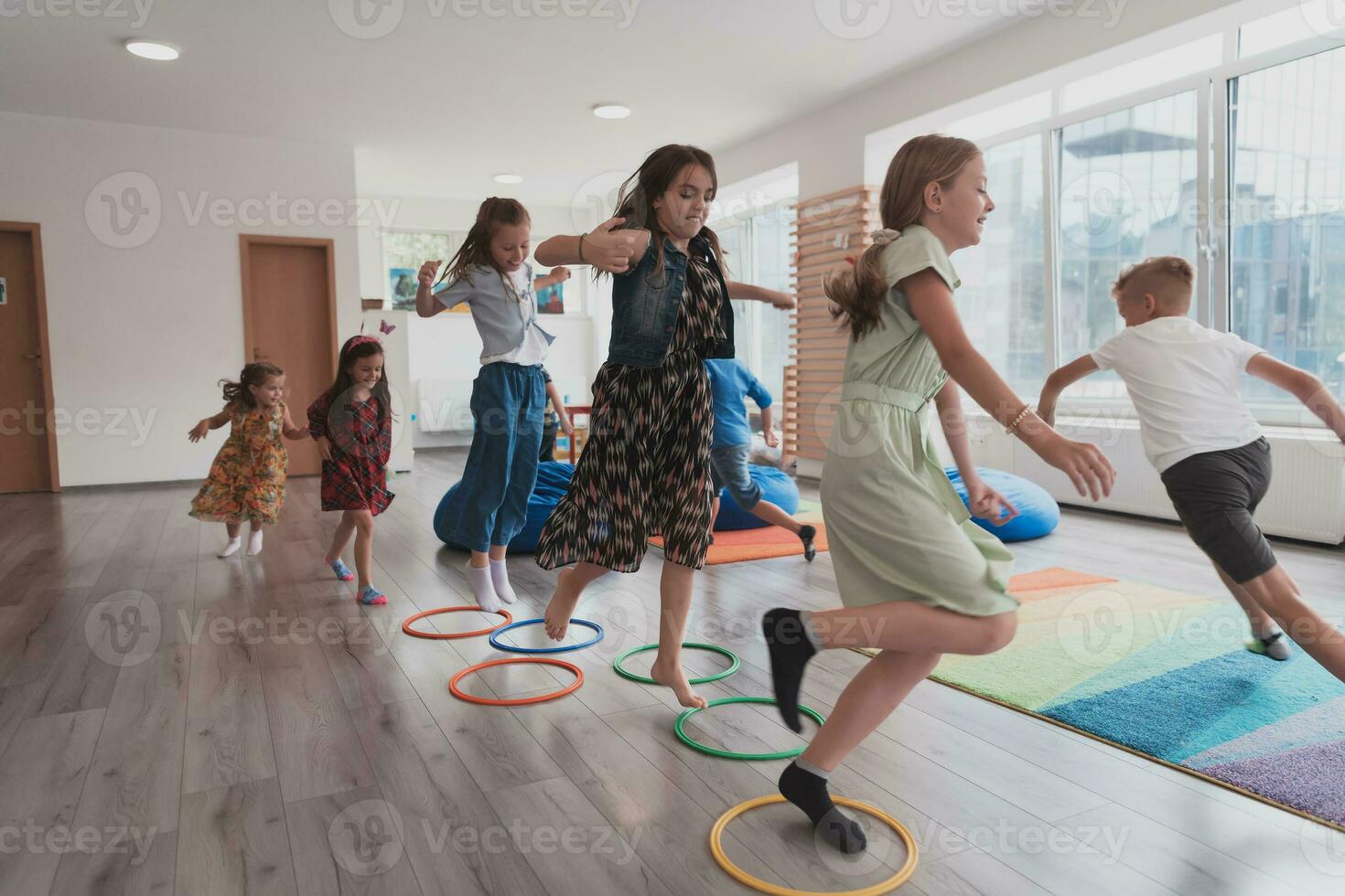 klein kinderkamer school- kinderen met vrouw leraar Aan verdieping binnenshuis in klas, aan het doen oefening. jumping over- hula hoepel cirkels bijhouden Aan de vloer. foto