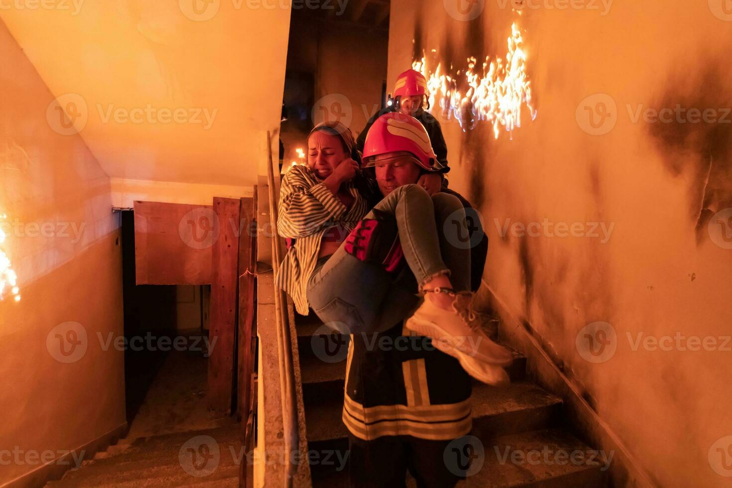 dapper brandweerman daalt af trap van een brandend gebouw en houdt opgeslagen meisje in zijn armen. Open brand en een brandweerman in de achtergrond. foto