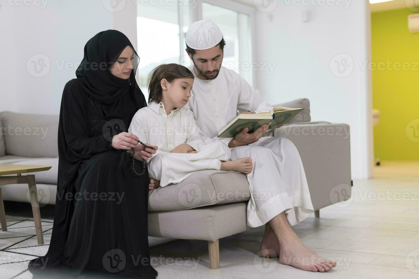 traditioneel moslim familie ouders met kinderen lezing koran en bidden samen Aan de sofa voordat iftar avondeten gedurende een Ramadan feest Bij huis foto