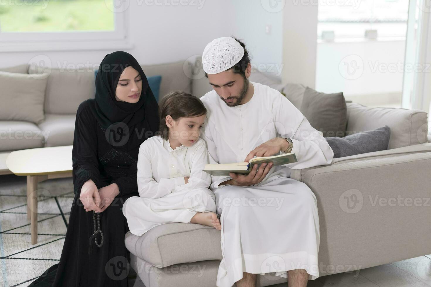 traditioneel moslim familie ouders met kinderen lezing koran en bidden samen Aan de sofa voordat iftar avondeten gedurende een Ramadan feest Bij huis foto