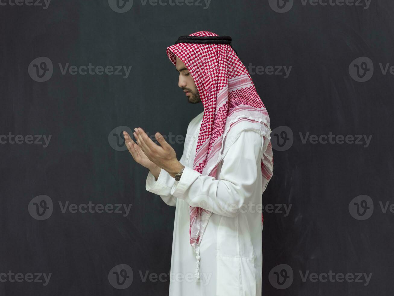 een jong Arabisch Mens in traditioneel kleren maken een traditioneel gebed naar god houdt zijn handen in bidden gebaar in voorkant van een zwart achtergrond foto