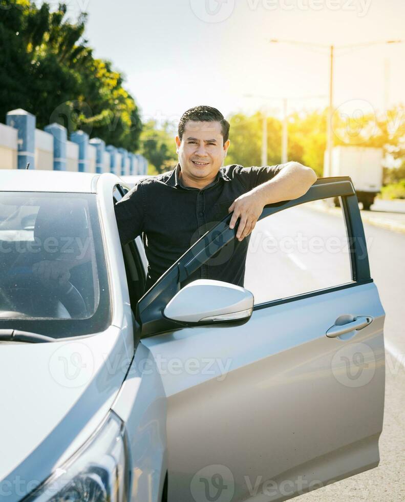 gelukkig bestuurder leunend Aan de auto deur in de straat, glimlachen bestuurder leunend Aan de auto deur. glimlachen eigenaar met nieuw auto. jong Latijns Mens leunend Aan auto deur in de straat foto