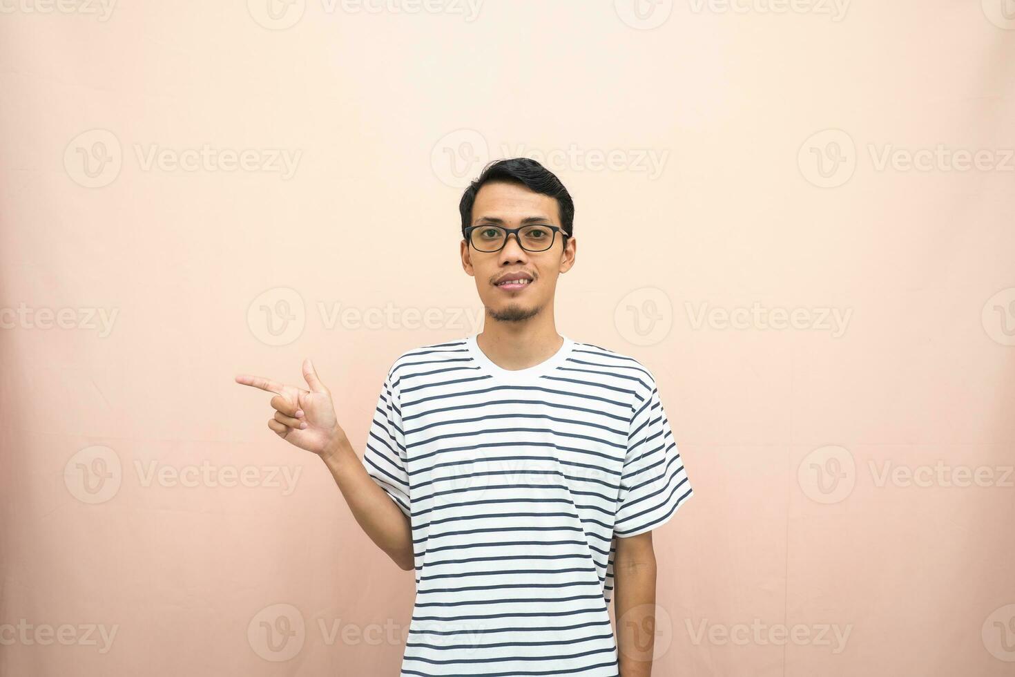 Aziatisch Mens met bril vervelend gewoontjes gestreept shirt, fluisteren houding terwijl richten naar de kant. geïsoleerd beige achtergrond. foto