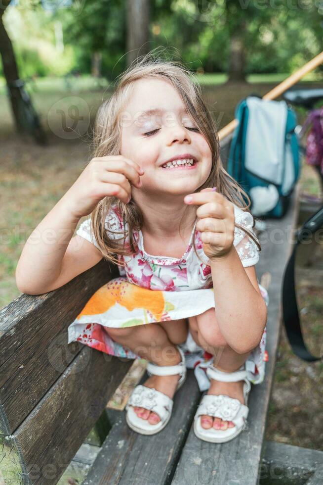 portret van schattig meisje met grappig uitdrukking zittend Aan een houten bank buitenshuis foto