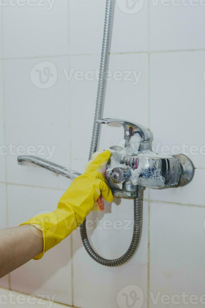 huisvrouw schoonmaak badkamer kraan en douche tik. hulp in de huishouding in geel beschermend handschoenen het wassen vuil bad tik. handen van vrouw het wassen bad foto