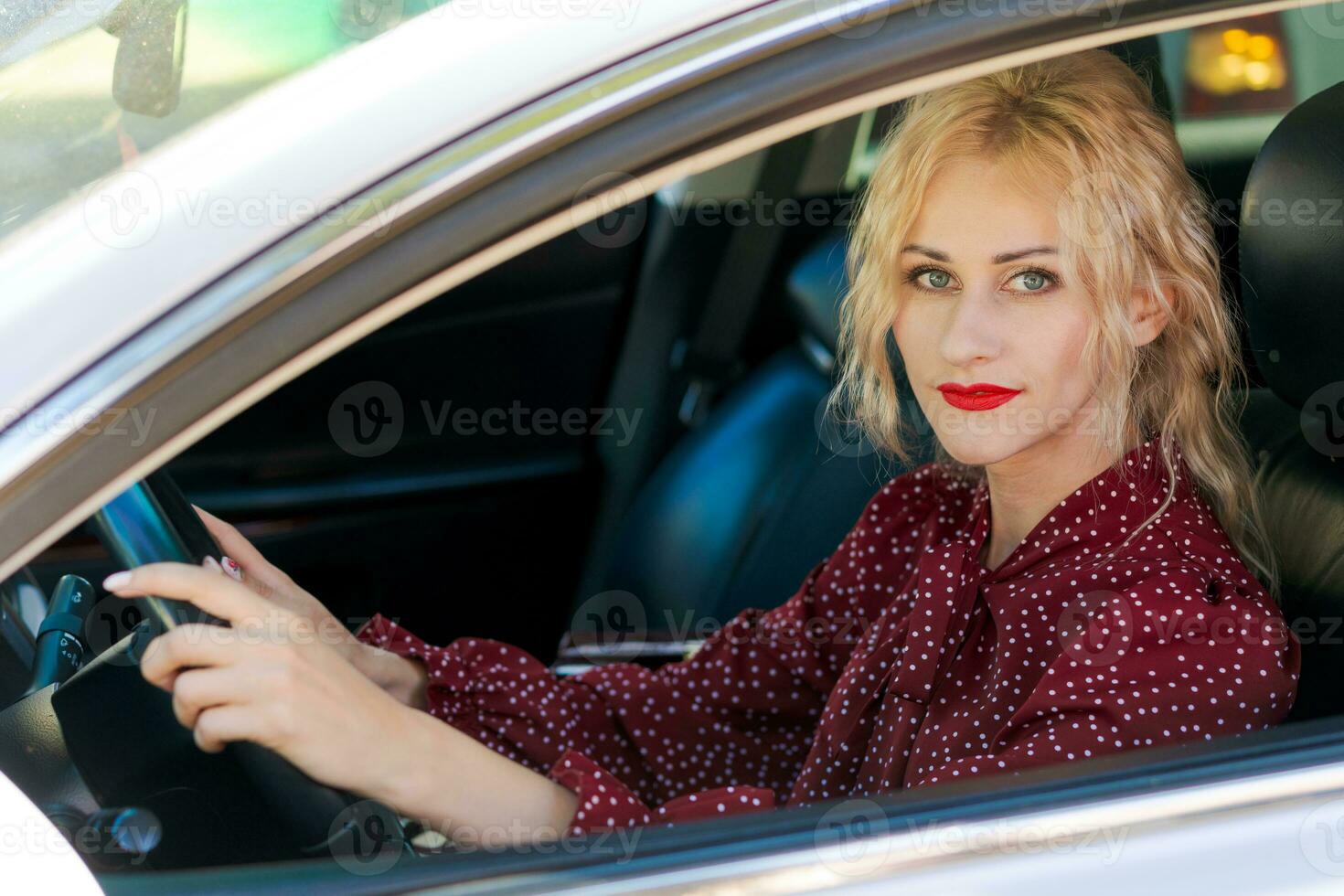 mooi geslaagd blond vrouw in een rood jurk zittend in een auto achter de wiel foto