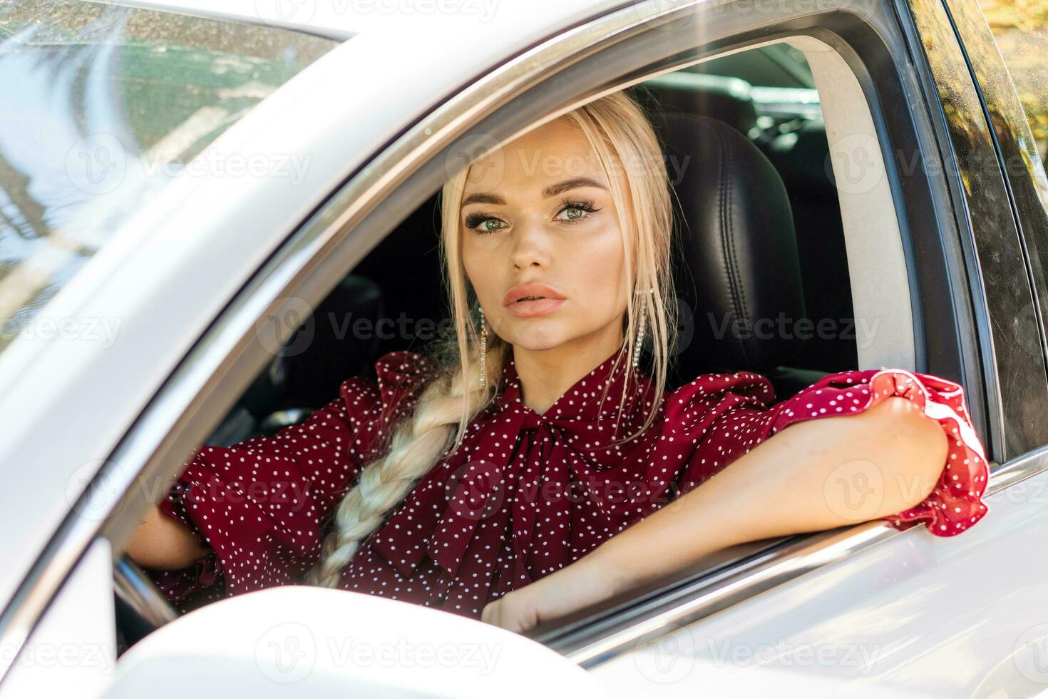 mooi geslaagd blond vrouw in een rood jurk zittend in een auto achter de wiel foto