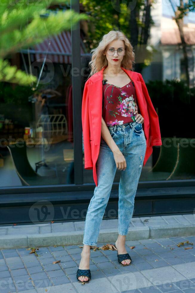 mooi blond vrouw in bril gekleed in rood jasje en blauw jeans poseren Aan straat in de stad foto
