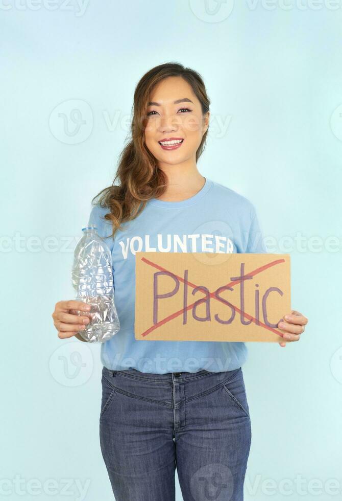 vrouw vrijwilliger in blauw t-shirt, handen houdt gebruikt plastic fles, anders hand- houdt karton teken Nee kunststof, op blauw achtergrond, concept campagne naar recycle gebruikt leeg plastic flessen, stop globaal opwarming foto