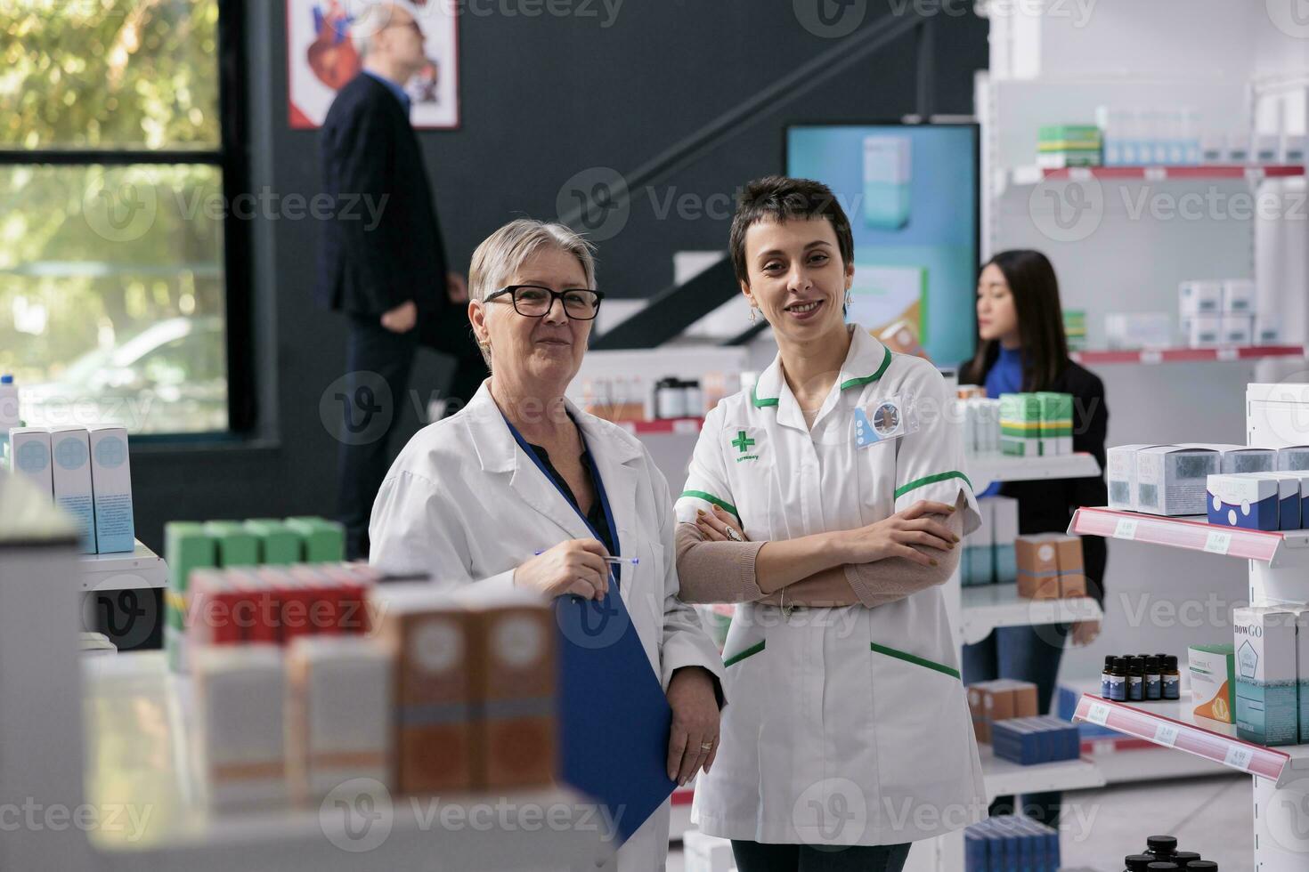 twee glimlachen farmaceutisch medewerkers staand in apotheek gangpad portret. jong en senior Dames apotheek specialisten vervelend apotheek medisch uniform op zoek Bij camera foto