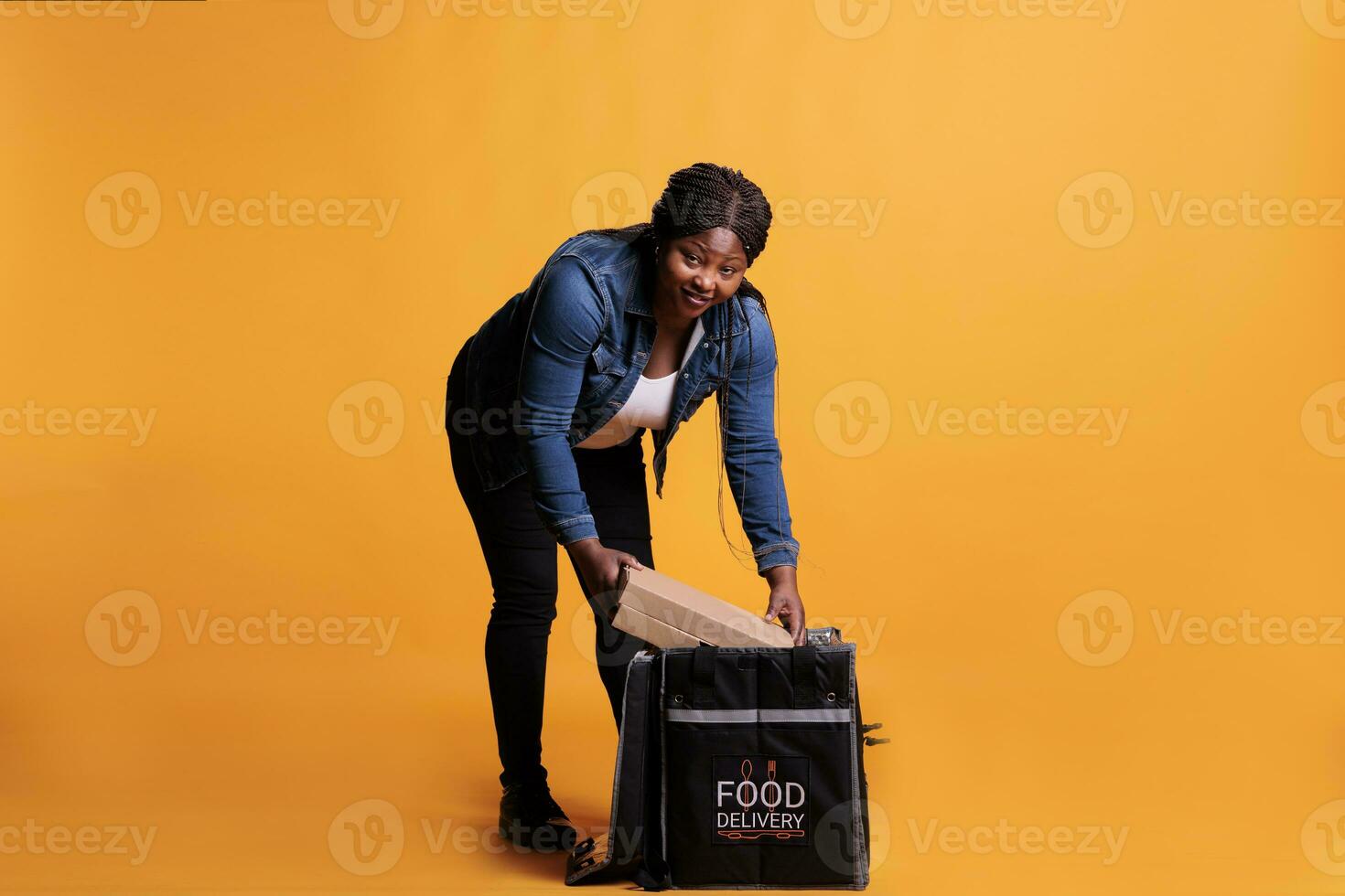 Afrikaanse Amerikaans werknemer Holding karton vol met pizza klaar naar leveren naar klant gedurende lunch tijd in studio met geel achtergrond. pizzeria koerier leveren meenemen voedsel maaltijd foto