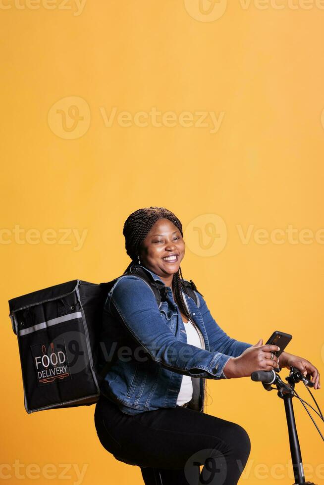 Afrikaanse Amerikaans koerier rijden fiets terwijl leveren meenemen voedsel maaltijd naar cliënt, controle adres Aan telefoon afhaalmaaltijd app, staand over- geel achtergrond in studio. nemen uit onderhoud en concept foto