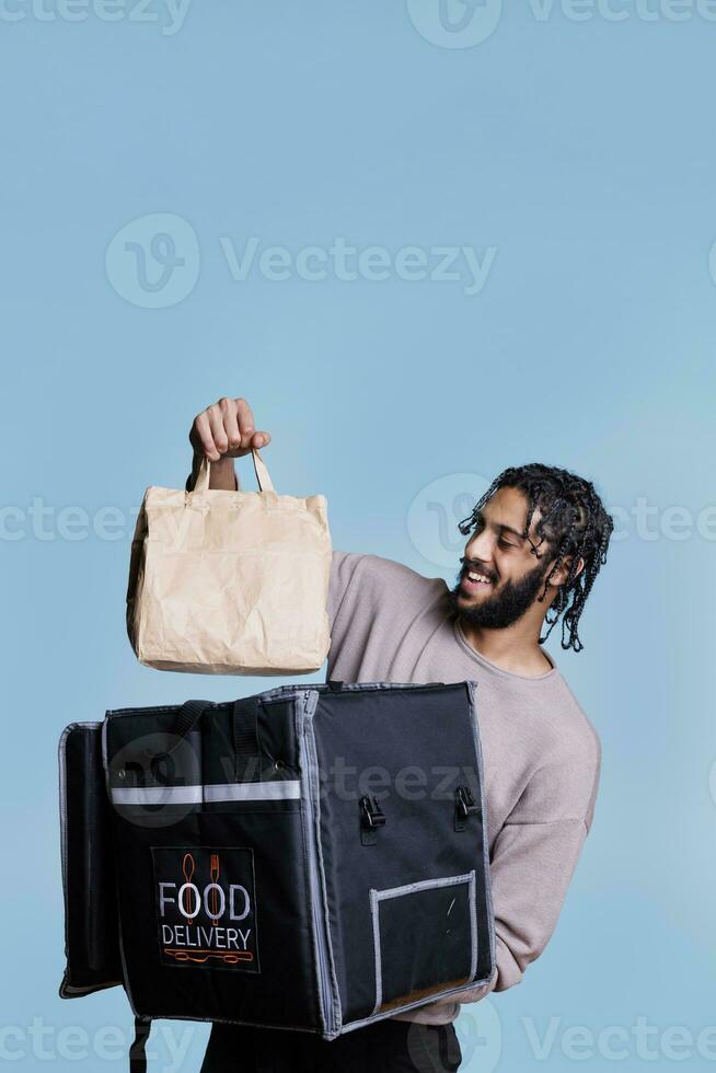 gelukkig Arabisch postbode Holding nemen uit papier zak terwijl leveren restaurant lunch. vrolijk voedsel levering onderhoud koerier met zorgeloos uitdrukking nemen pakket van thermisch rugzak foto