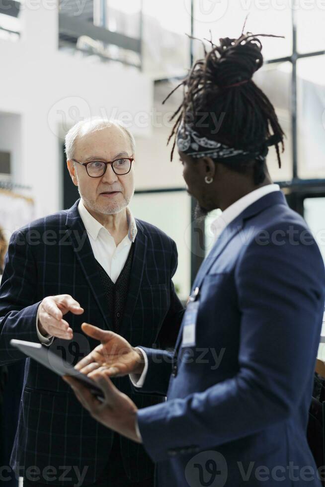 Afrikaanse Amerikaans werknemer controle overhemd voorraad Aan tablet computer voor ouderen klant in boodschappen doen centrum. senior cliënt buying formeel kleren en modieus accessoires in modern winkel foto