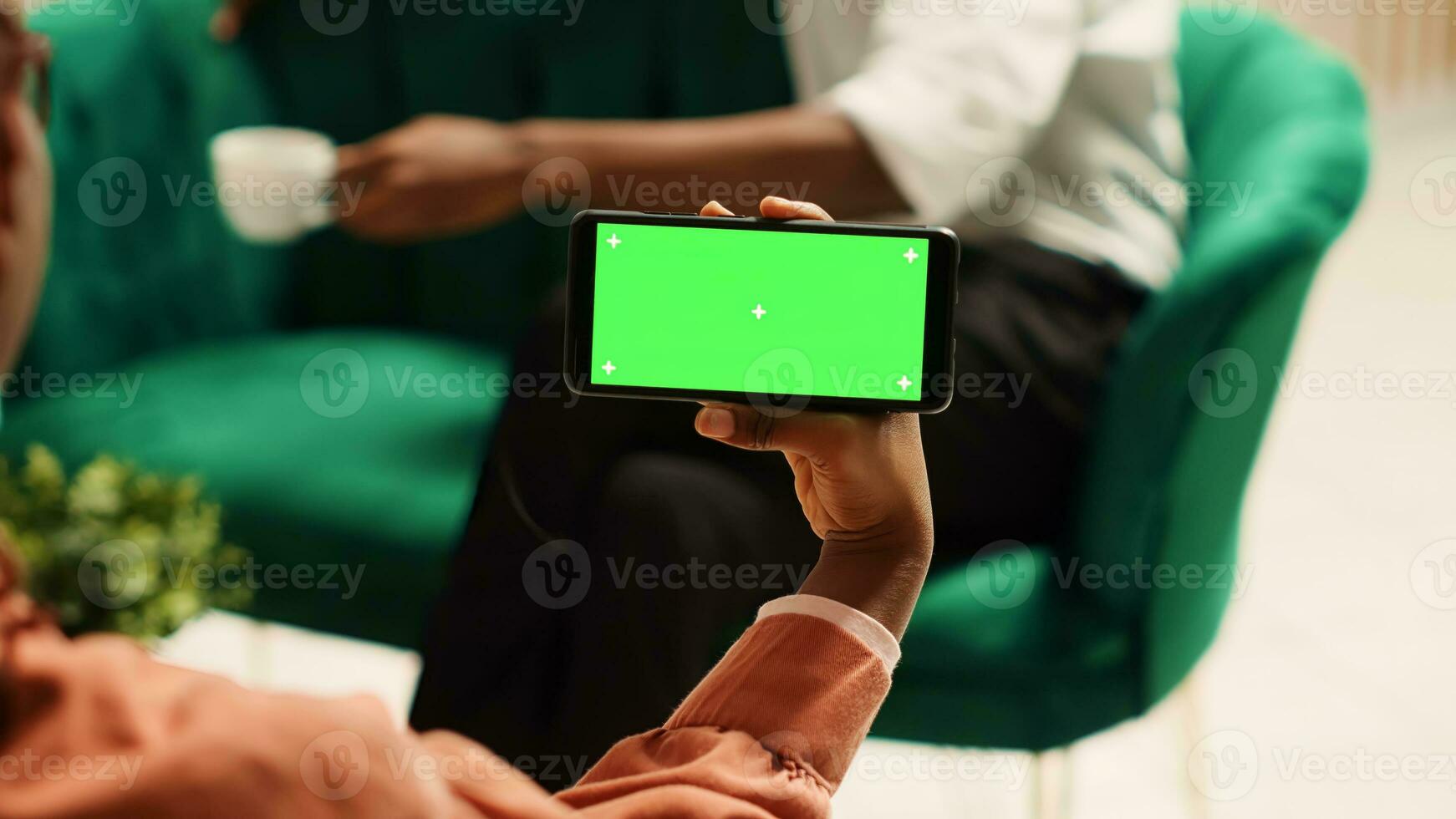 Afrikaanse Amerikaans vrouw Holding chroma sleutel groen scherm bespotten omhoog smartphone in landschap mode zittend Aan knus bank. toeristen aan het wachten in elegant hotel lounge naar worden gecontroleerd in foto